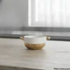 Ceramic soup bowl with gold handles on wooden table near window